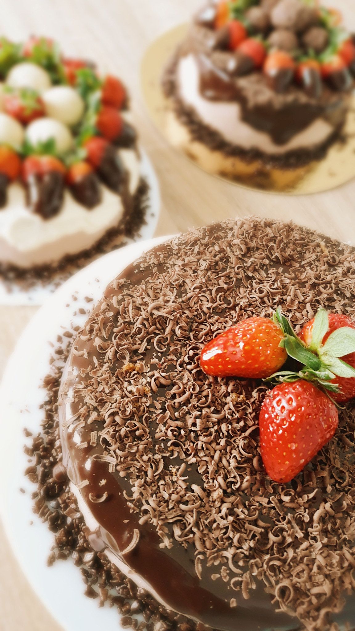 Torta de chocolate coberta com raspas de chocolate e morangos, com outras tortas ao fundo