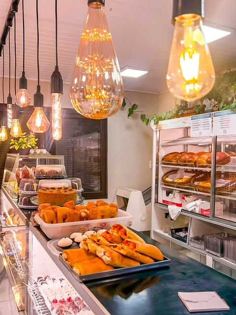 Interior da Atalibio Confeitaria com luminárias de lâmpada, prateleiras de pães e vitrine de salgados
